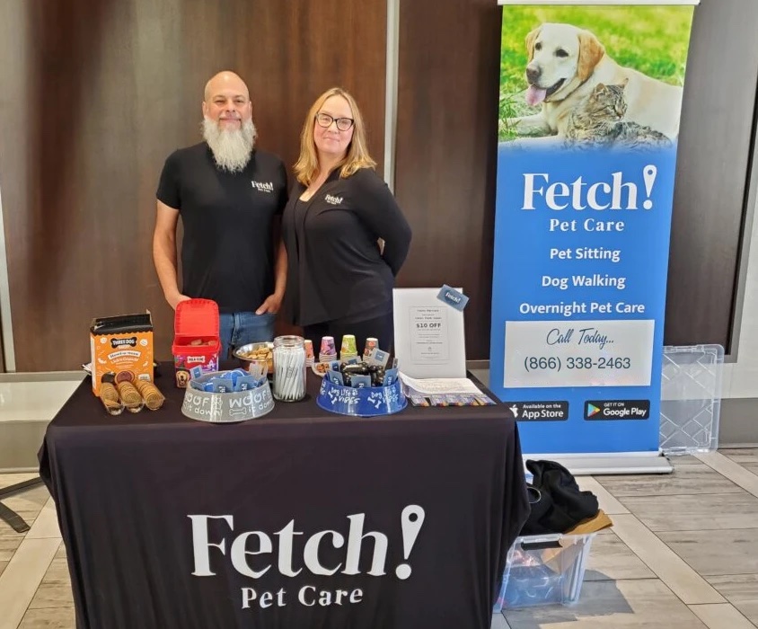 Two Fetch! Pet Care team members standing behind a branded table of pet treats and giveaways next to a Fetch! banner